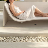 A woman relaxing on a modern, curved lounge chair in a minimalist setting with pebbles in the foreground.