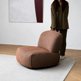 Brown chenille armless swivel chair on a light rug, barefoot person standing beside it in a bright room with artwork