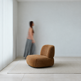 Brown upholstered armless swivel chair on a light rug in a bright empty room, blurred woman walking past