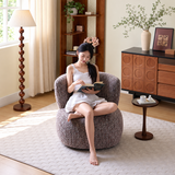 Woman reading in a brown textured loop yarn swivel barrel chair with a cream rug and wooden cabinet in a sunlit living room