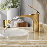 Brushed gold waterfall faucet pouring water into a beige marble sink, with a candle and plant in the background.