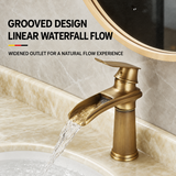 Brushed gold faucet with a widened grooved waterfall spout, flowing water over a beige marble basin.