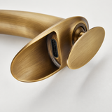 Close-up detail of a brushed brass kitchen faucet's open waterfall spout and round handle against a white background.