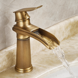 Bronze kitchen faucet with waterfall-style spout, water flowing gracefully from the angled outlet, mounted on a marble sink.