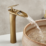Brushed brass waterfall faucet with flat lever handle, water pouring into a ridged vessel sink.