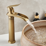 Brushed brass tall kitchen faucet with flat lever handle, water flowing into a ribbed basin beside soap dispensers.