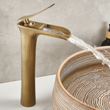 Brushed brass tall single-handle faucet beside a wood-grain vessel sink, water arcing from the open-channel spout.