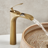 Brushed gold single-handle faucet pouring water into a wood-grain round basin against a gray marble wall.