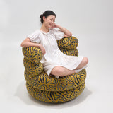 Woman seated cross-legged on a mustard barrel chair with black squiggle-pattern upholstery in a white studio