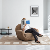 Brown textured swivel barrel chair in a white room, person reading while seated, geometric rug and framed print behind