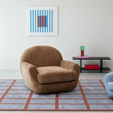 Brown textured swivel barrel chair on a blue and rust grid rug in a bright room with a black low shelf and wall art