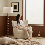 Woman seated in a cream boucle swivel barrel chair by a shuttered window and floor lamp.