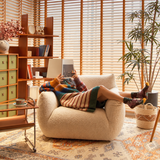 Woman with legs resting on a cream bouclé barrel chair in a warm room with wooden blinds