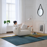 Woman reclining in a cream bouclé swivel barrel chair in a bright white paneled room with a blue-green rug