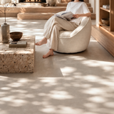 Cream bouclé swivel barrel chair with a seated reader, travertine table and wooden steps in sunlight