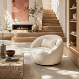 Cream bouclé swivel barrel chair with pillow on a sunlit floor, wooden stairs, fireplace and stone table beside it