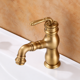 Brushed antique brass kitchen faucet with a carved lever handle and curved spout, shown on a white sink against a tan wall.