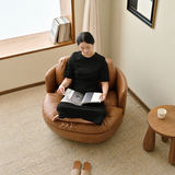 Woman reading in a brown leather-look swivel barrel chair on a textured rug, high-angle view in a bright room