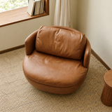 Tan leather-look swivel barrel chair on a beige pebble rug beside a wooden ledge and cream curtain