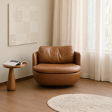 Tan leather-look swivel barrel chair by sheer curtains, wood pedestal table and abstract wall art