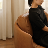 Woman in black seated in a brown leather swivel barrel chair by sheer curtains