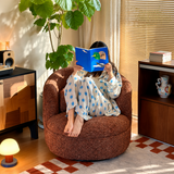 Rust-brown bouclé swivel barrel chair with a person reading a blue book, sunlit room with plant and blinds