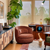 Rust barrel swivel chair beside a wooden record console, with an orange cat walking across a patterned rug
