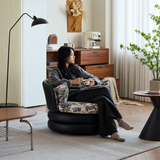 Woman with a mug relaxing in a black swivel barrel chair with toile cushions beside a wood side table and floor lamp