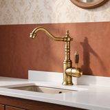 Brass kitchen faucet with wood-grain handle and curved spout, on a white countertop with a sink, against a warm wall.