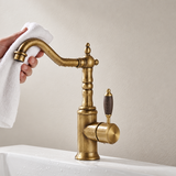 Brass kitchen faucet with wood-grain handle and curved spout, positioned over a white sink with visible water droplets.