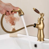 Hand pulling out a brushed brass faucet spray head, water flowing, wood-grain handle, white basin.