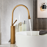 Antique brushed brass kitchen faucet with a curved spout pouring water into a white vessel sink beside two elegant bottles.