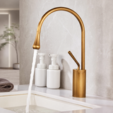 Antique brushed brass kitchen faucet pouring water into a white vessel sink beside two white bottles.