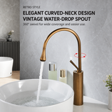 Antique brushed brass faucet with curved neck and red swivel arrows, pouring into a white basin against a marble wall.