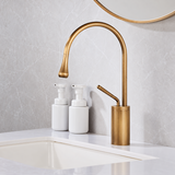 Antique brushed brass high-arc kitchen faucet with side lever beside a white vessel sink and two white bottles.