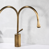 Antique brushed brass kitchen faucet with a curved dual spout and a side lever handle against a light gray marble background.