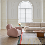 Dusty pink velvet swivel barrel chair angled in a light paneled room with a bouclé sofa, glass lamp table, and patterned rug.